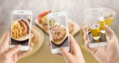 Three people taking photos of food, one displays sausage and fries, another a steak and the last one a beer.