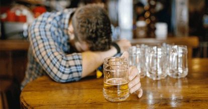 A man with his head in his arms at a bar table with empty mugs around him and a beer in his hand