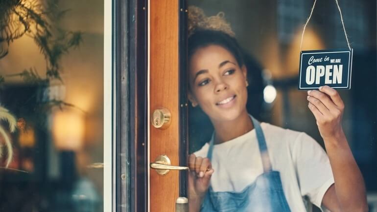 Women working at a restaurant smiles and holds a sign that says the venue is open.