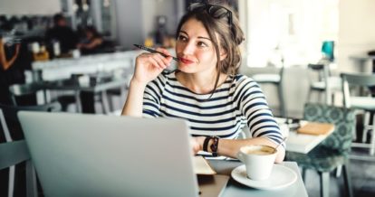 woman works on laptop in restaurant while holding pen in mouth thinking