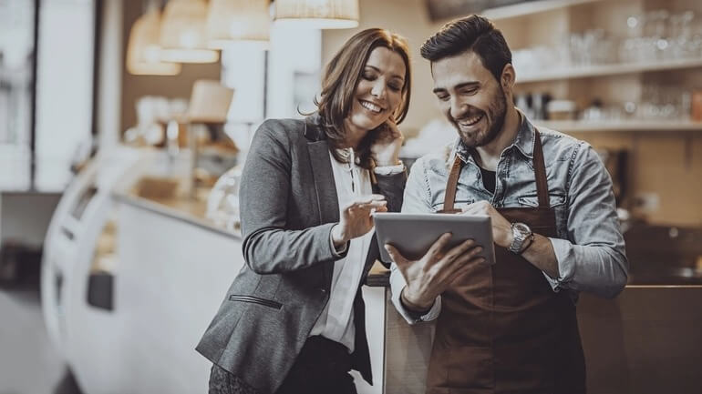 A female manager trains a new male employee while looking at a handheld tablet in a restaurant.