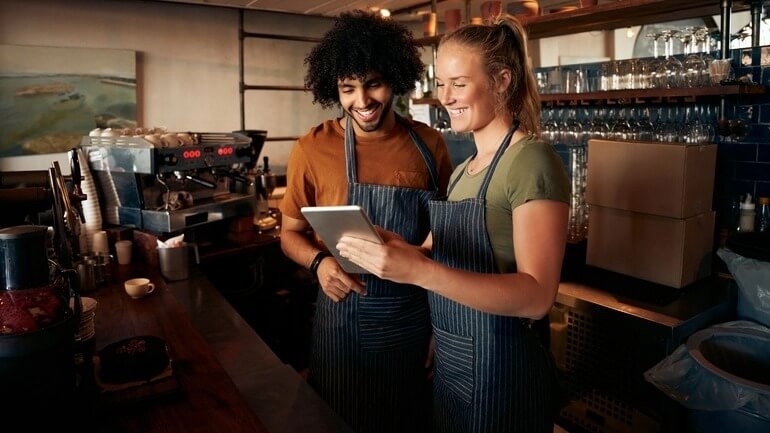 A male and female coworker read from a handheld tablet together while smiling in a restaurant.