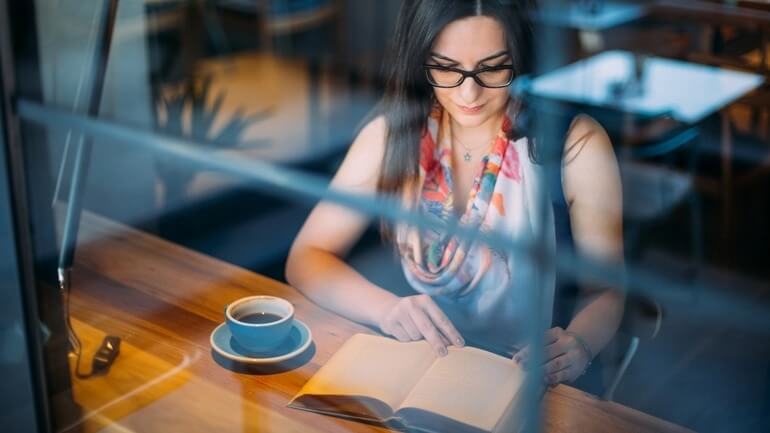 A woman reads a book in a restaurant with a cup of coffee next to her