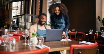 Two smiling restaurant workers using a laptop in an empty restaurant.