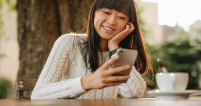 A woman looks at her phone while enjoying coffee outside at a table.