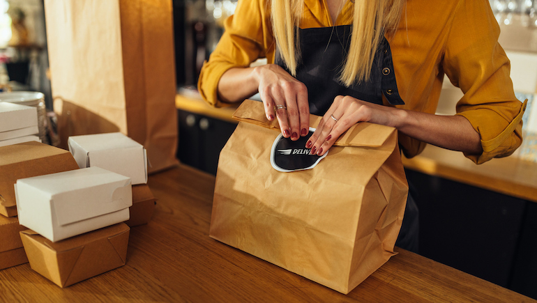 Close up of young woman packing food in paper bags and preparing them for delivery.