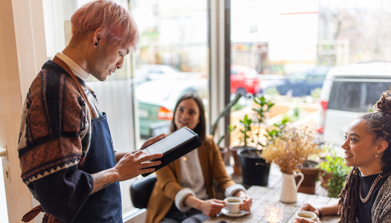 Man taking orders from women sitting in the cafe. Male waiter wearing apron holding digital tablet taking orders from female customers.