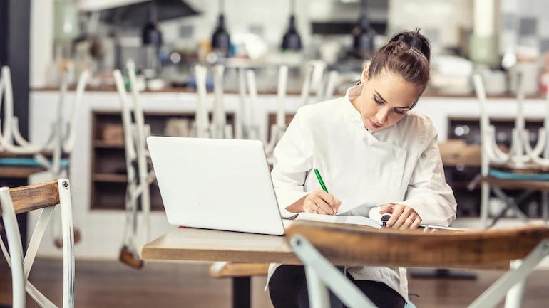 Female restaurant chef sits at the table after hours doing her bookkeeping in front of an open laptop.