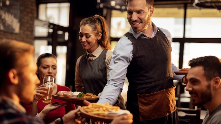 Happy waiters bringing food at the table and serving group of friends in a restaurant.