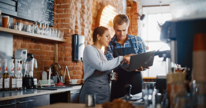 Two Diverse Entrepreneurs Have a Team Meeting in Their Stylish Coffee Shop. Barista and Cafe Owner Discuss Work Schedule and Menu on Laptop Computer. Multiethnic Female and Male Restaurant Employees.