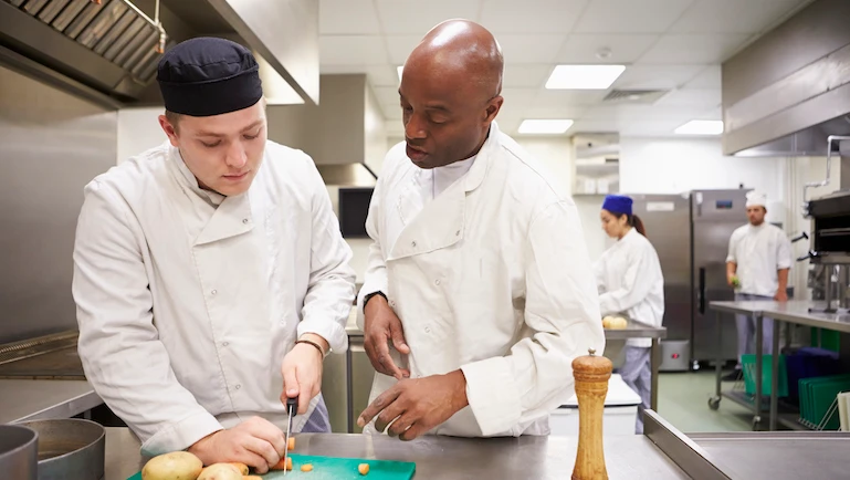 Teacher Helping Students Training To Work In Catering Chopping Vegetables