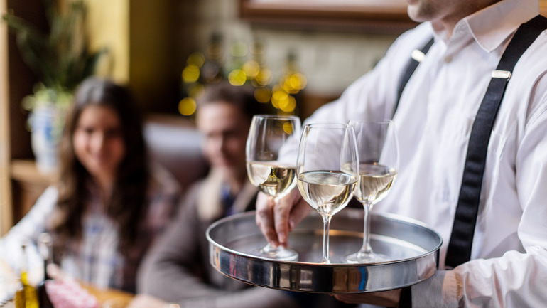 Man wearing white dress shirt holding three glasses of wine on a silver tray.