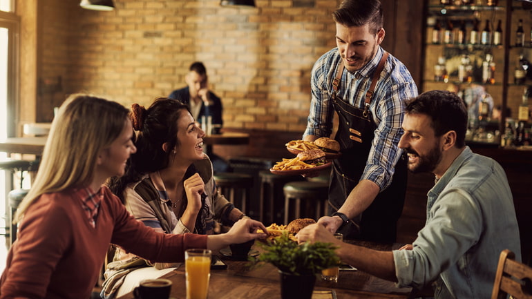 A man who is a server brings a group of three happy customers their burgers and fries.