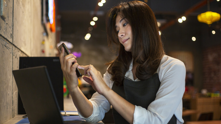 A woman wearing white and black is sitting in front of a laptop on her mobile phone in a restaurant.