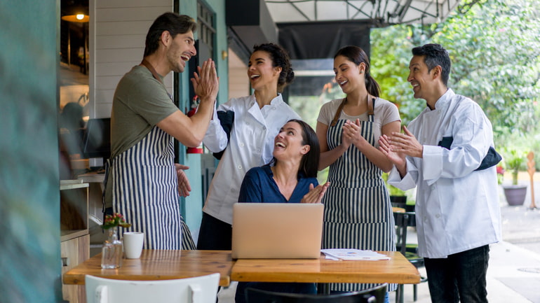 Five restaurant workers outside of their restaurant gathered around a table, having a meeting clapping.