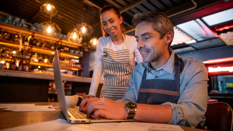 Man sitting in a restaurant doing work on a computer while a woman is standing behind him helping.