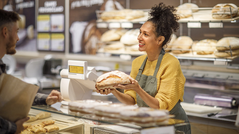 Worker at a bakery is holding a loaf of bread to show to a customer.