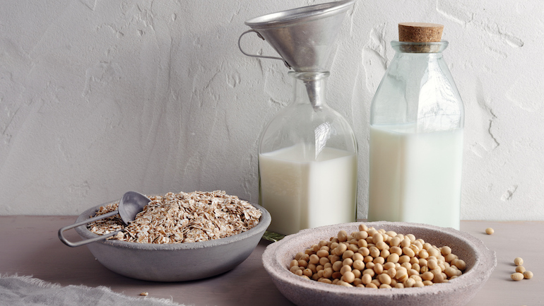 Glass jugs of soy and oat milk with bowls of soybeans and oat flakes.