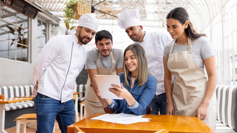 A group of restaurant staff standing and gathering around another staff member sitting looking a tablet.