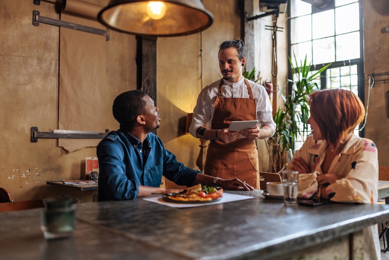 A waiter wearing an apron taking an order from a man and woman sitting in a restaurant.